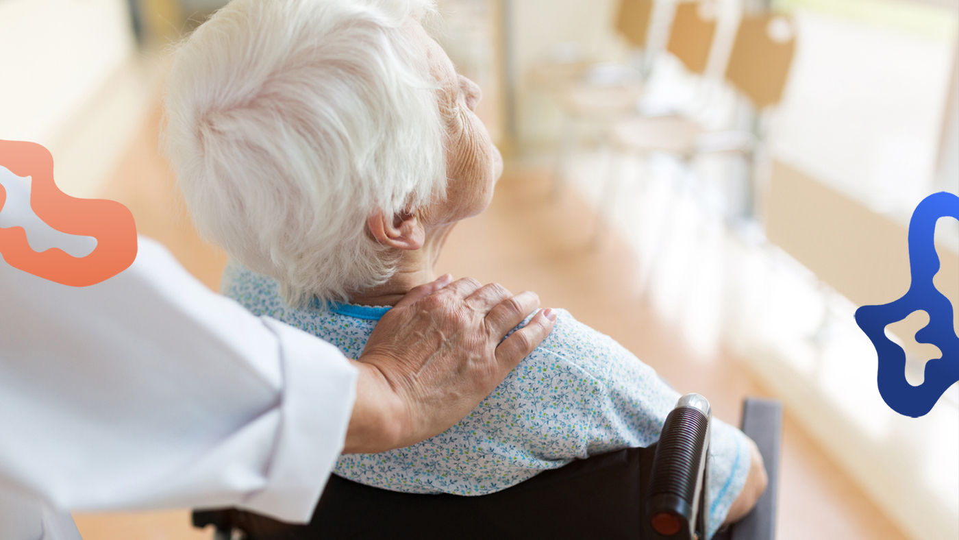 En undersköterska lägger en trygg hand på axeln på en äldre patient som sitter i rullstol.